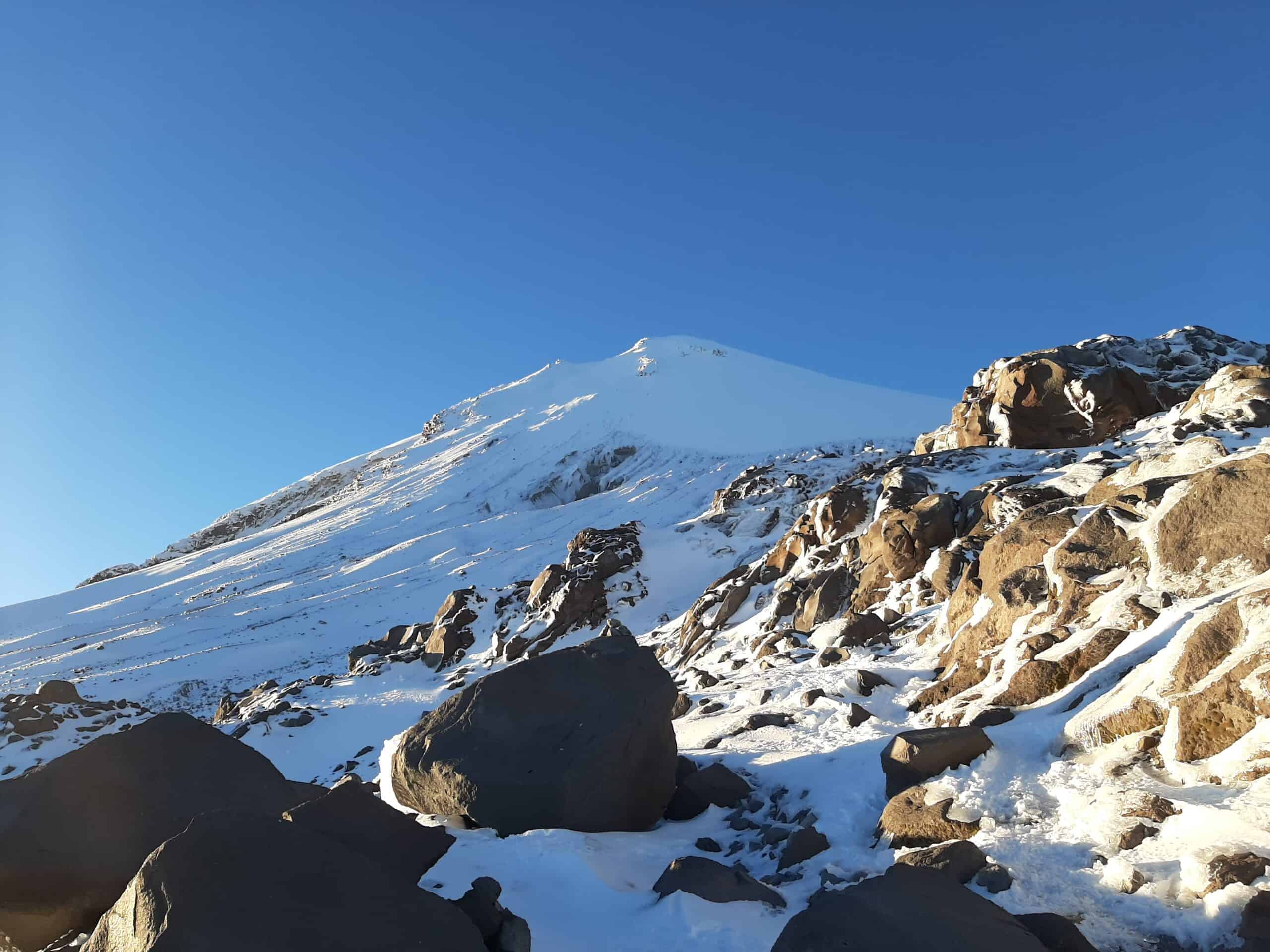 Snow-covered upper slopes of Pico de Orizaba with volcanic rock outcrops in the foreground under a clear blue sky