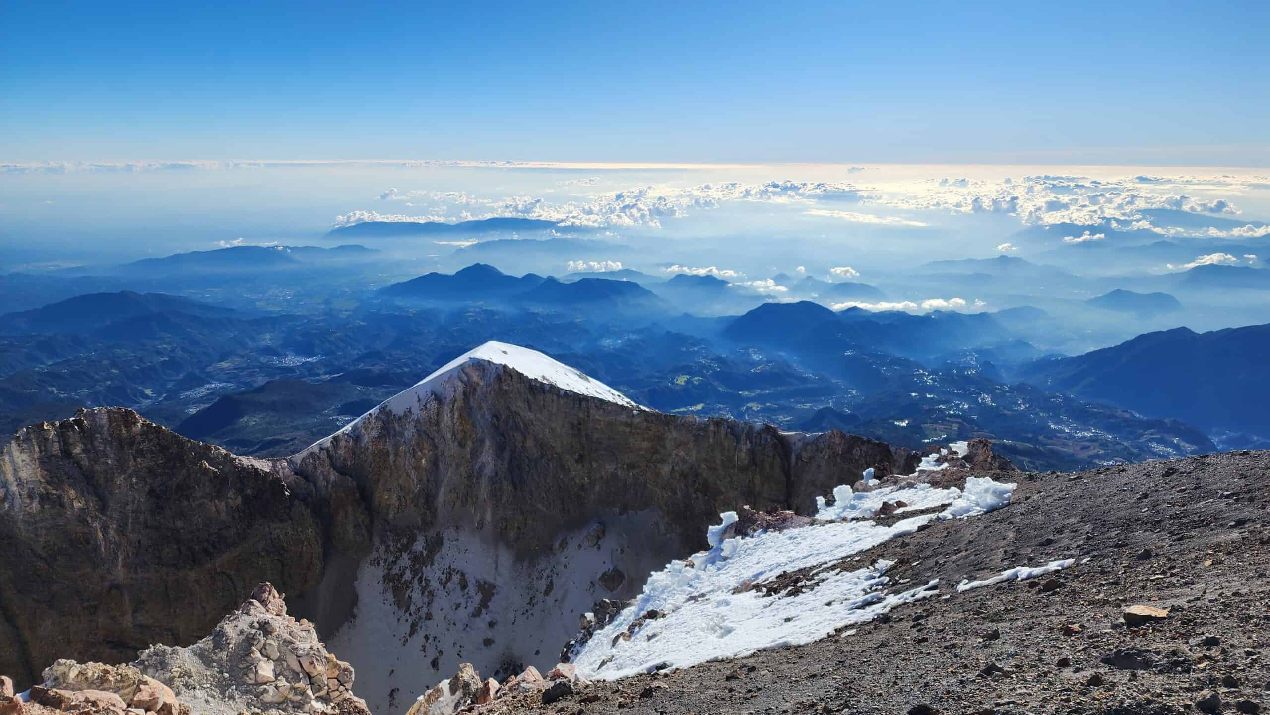 View from the crater rim of Pico de Orizaba looking down into the volcanic crater with layered mountain ranges and clouds stretching to the horizon