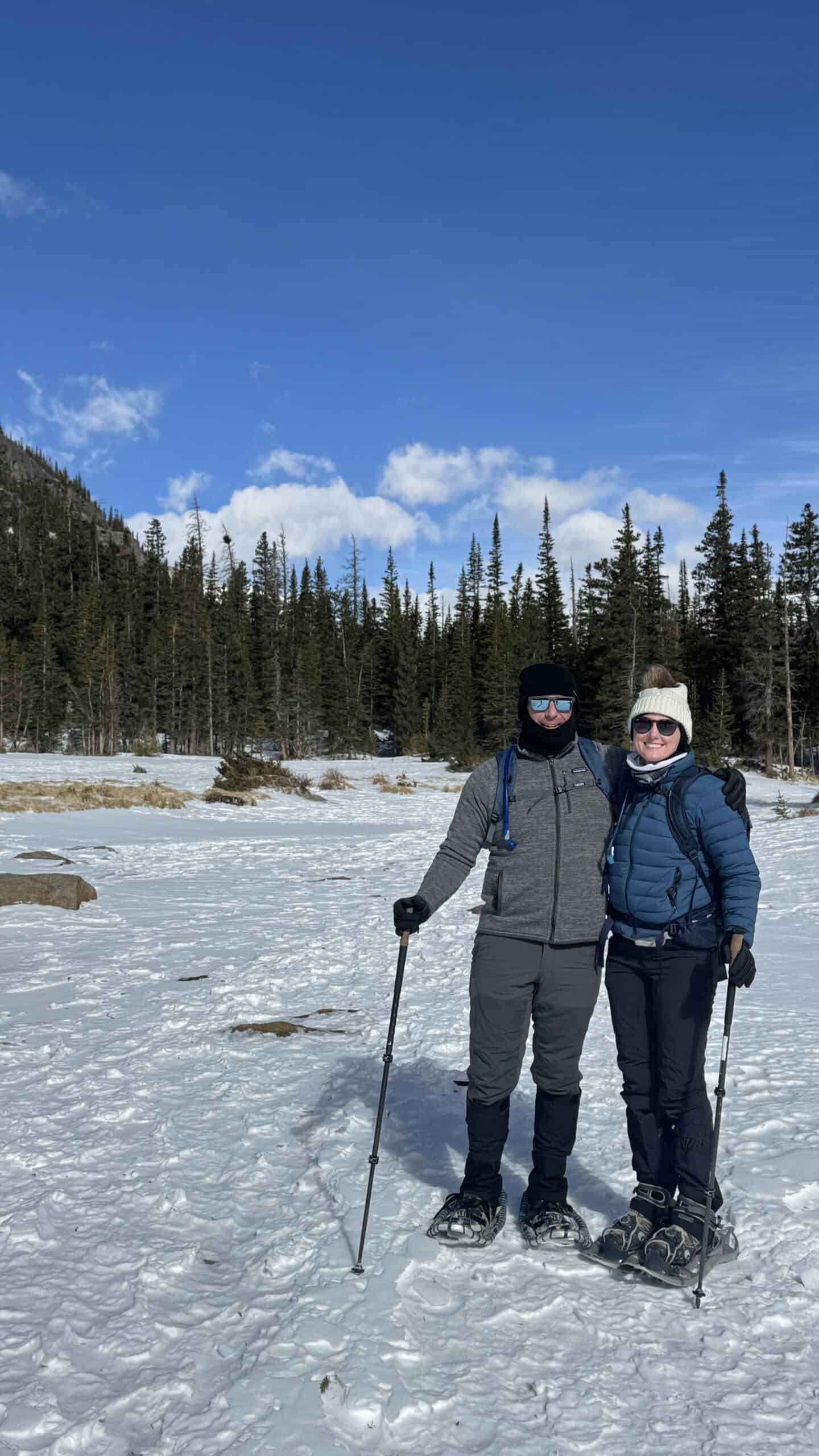 Couple snowshoeing in Rocky Mountain National Park, standing arm in arm on a snowy meadow with a forest of pines and a mountain ridge under a bright blue sky behind them.