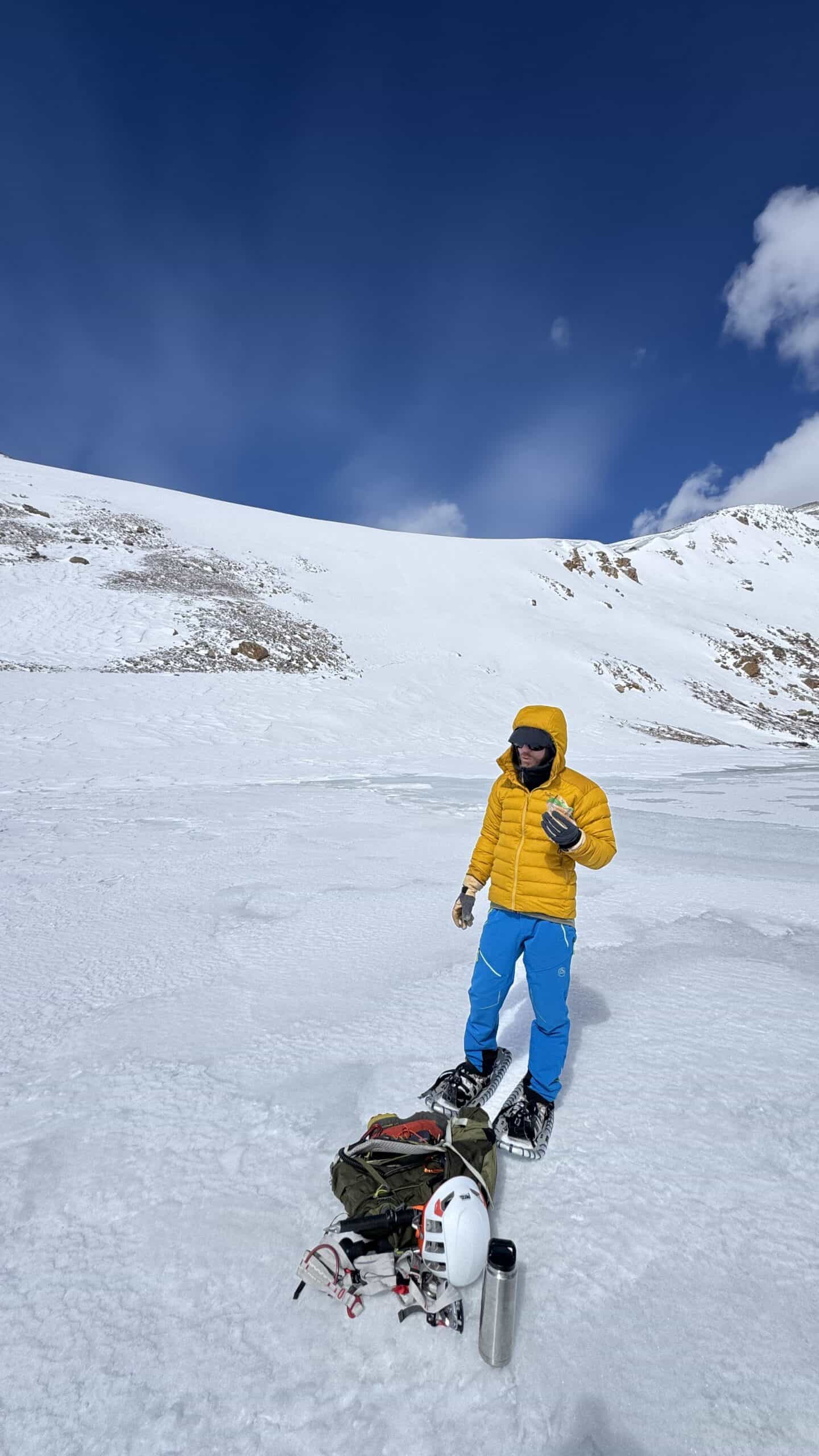 Snowshoer in a yellow down jacket standing on a frozen alpine lake next to a pile of climbing gear, helmet, and thermos, with a snow bowl rising behind him.