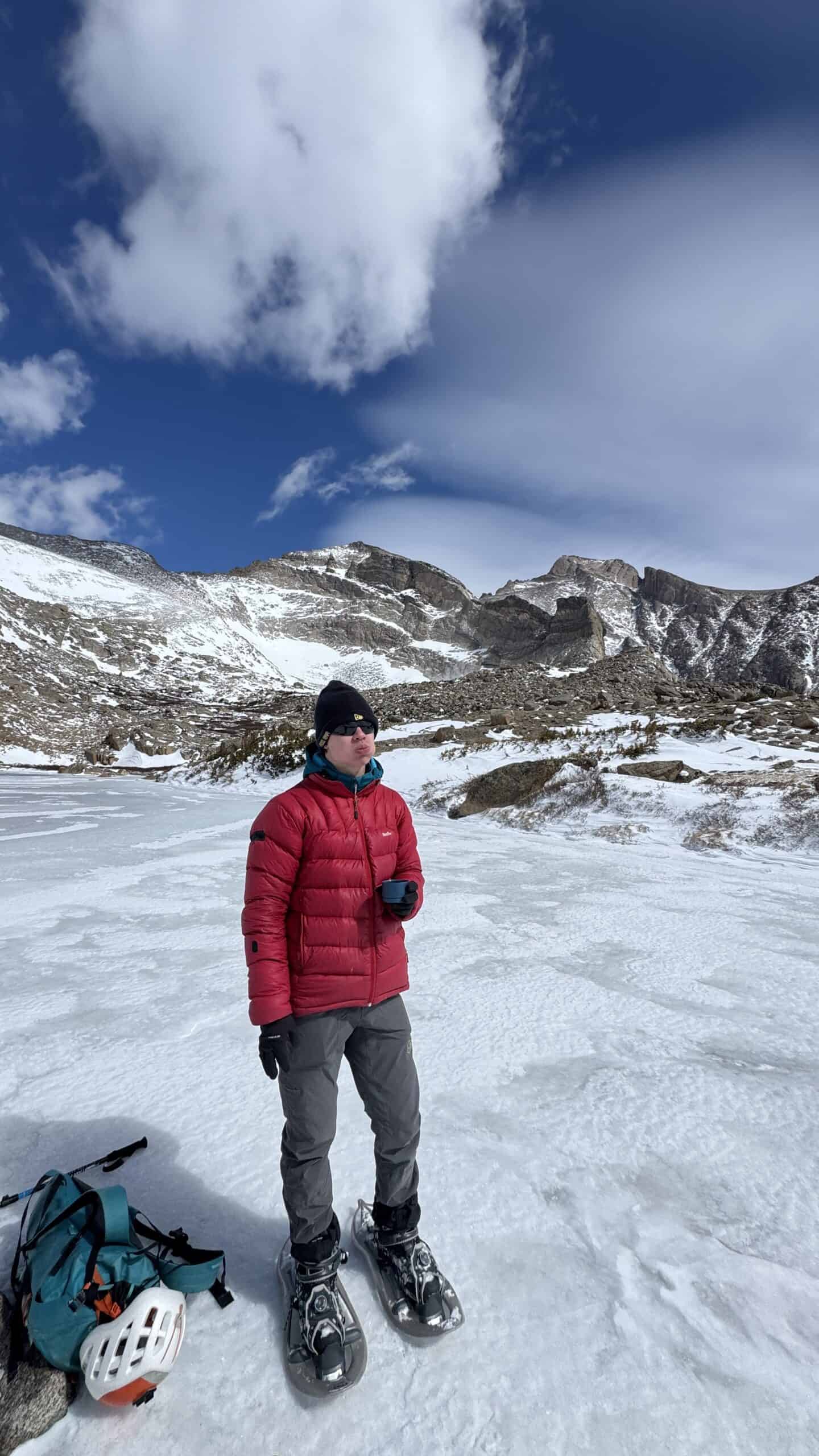Snowshoer in a red down jacket sipping a hot drink on a frozen alpine lake, rocky snow-covered peaks and a large cloud behind him.