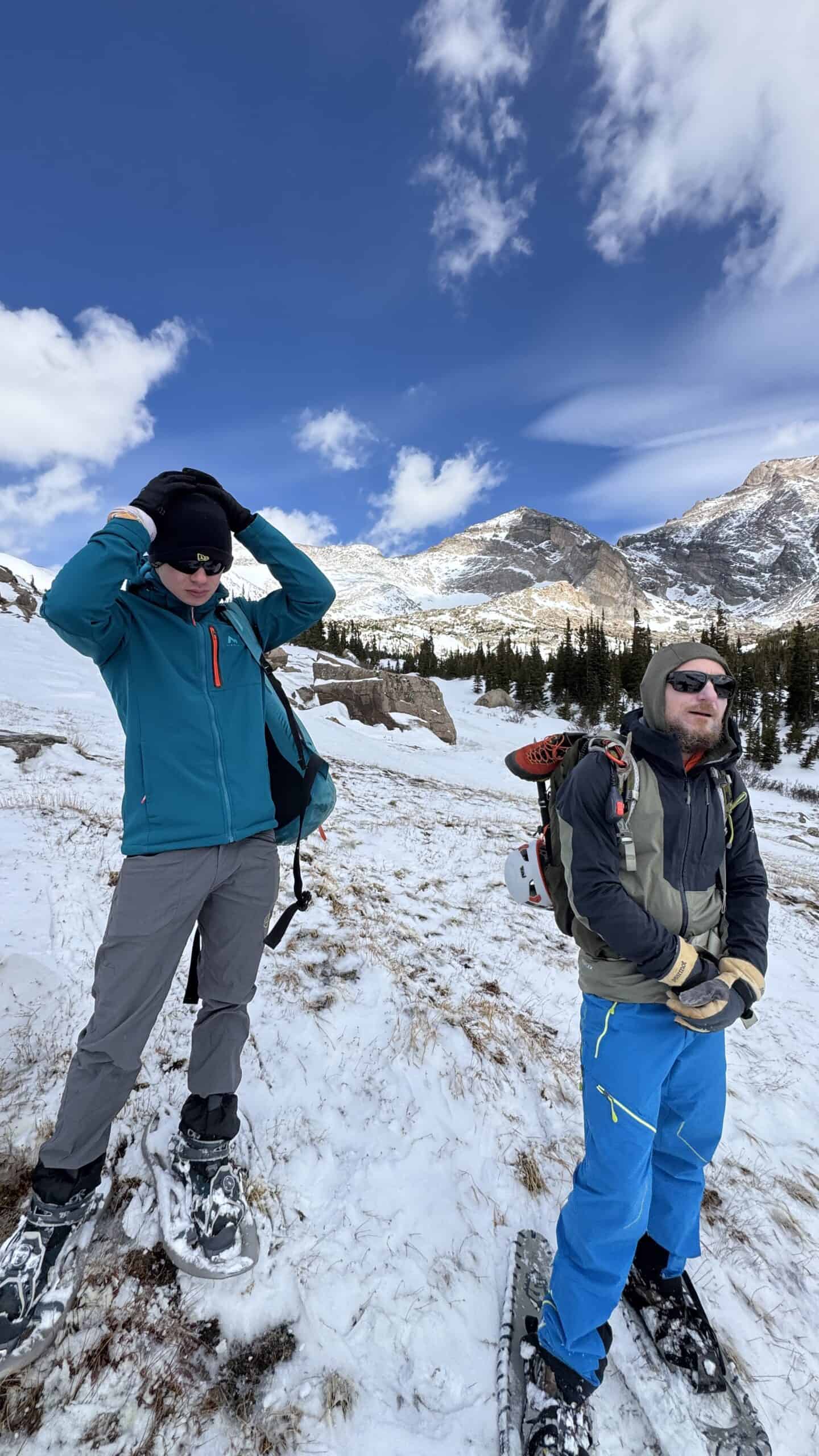Two snowshoers pausing on a grassy, snow-patched slope with rugged mountain peaks and evergreen forest behind them.