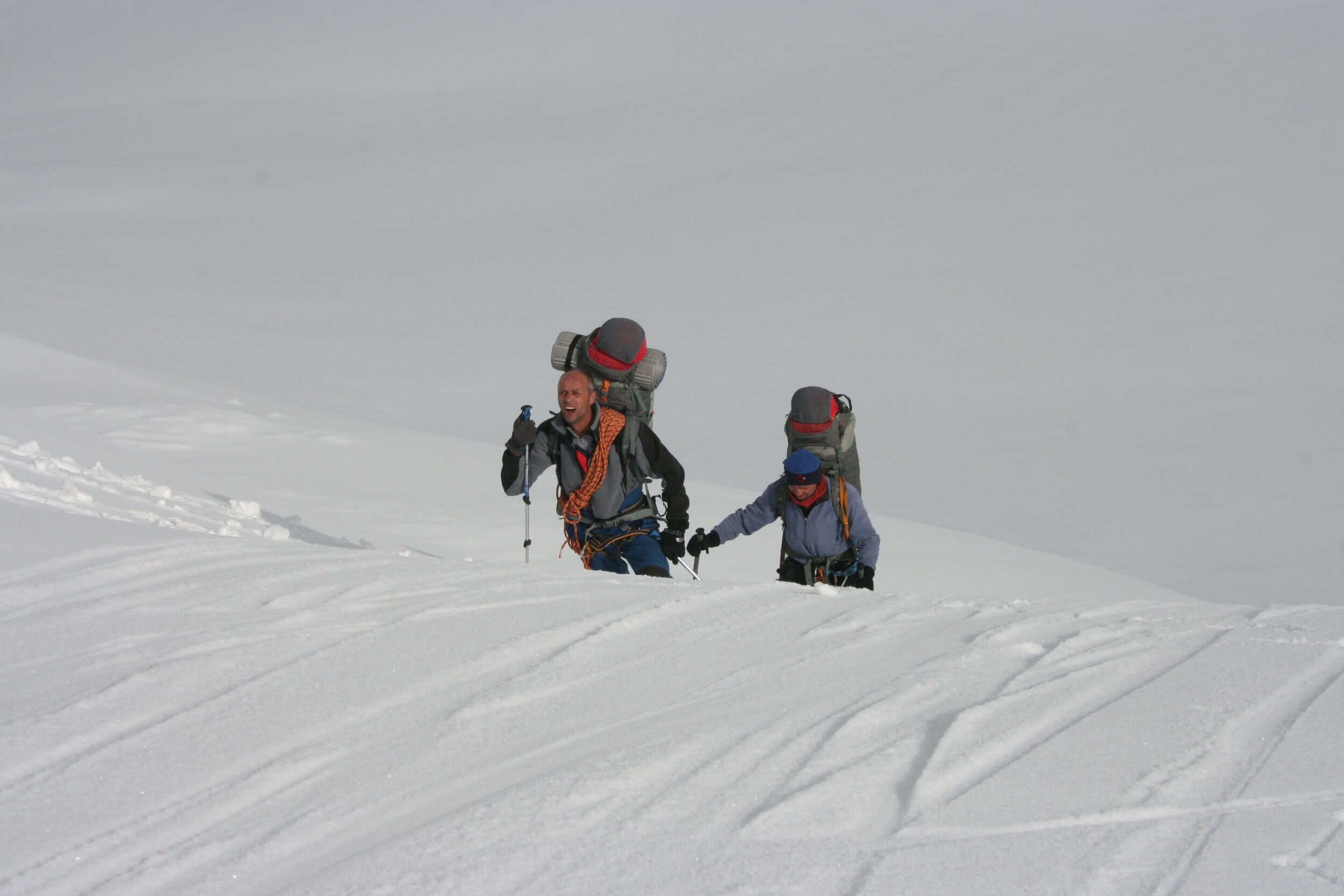 Two roped climbers with heavy packs pushing uphill through flat light and deep snow during Alpine Pro-1